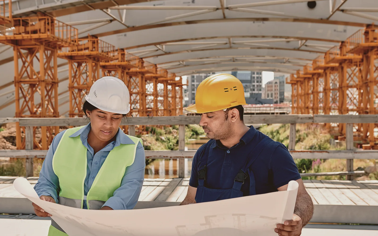 Two contractors lean over large printed construction drawings spread across a folding table at a job site, one marking a detail with a pen while the other points at the same sheet