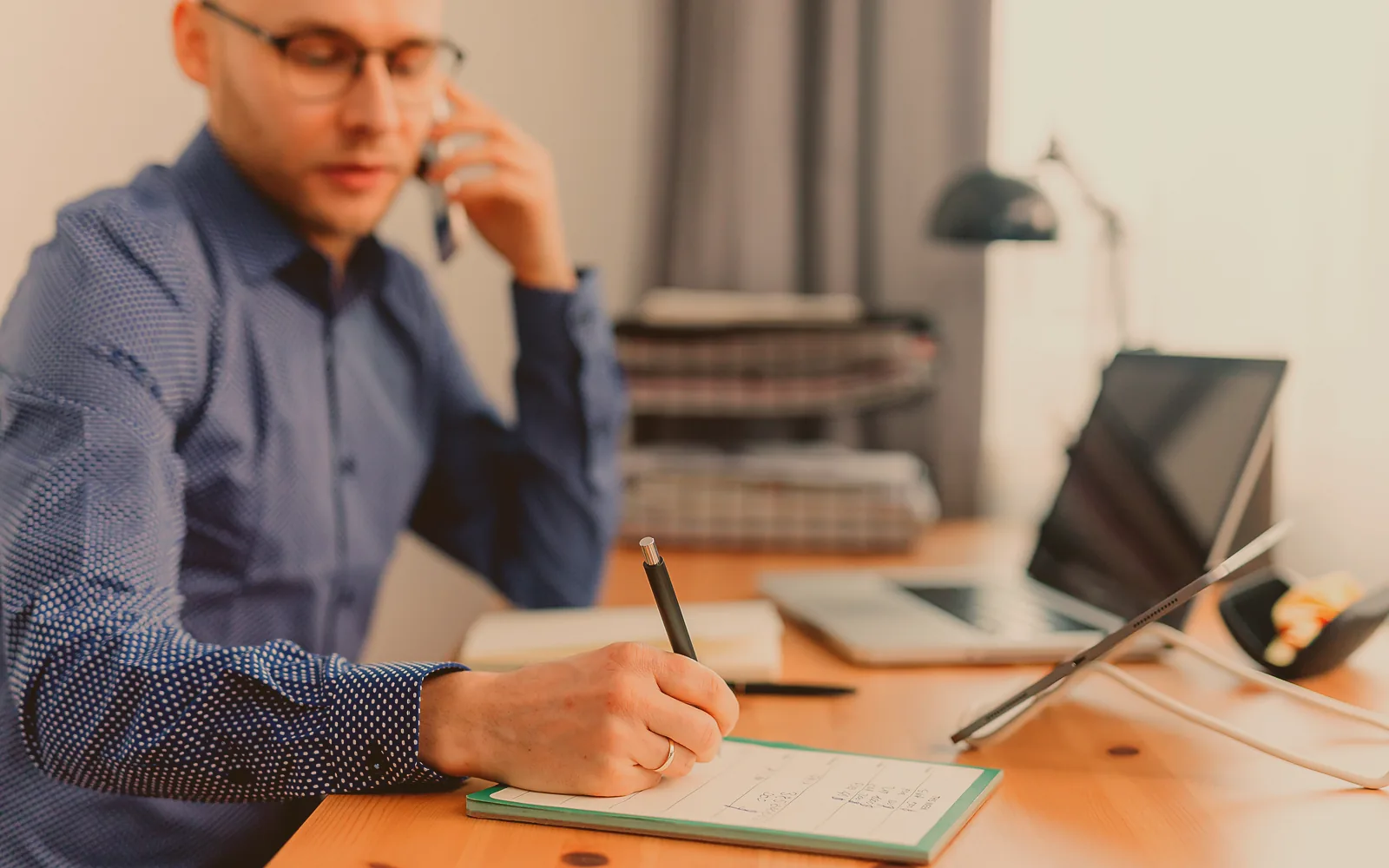 An office manager holds a landline handset against her shoulder while she types a new caller's details, a second phone and a printed job sheet beside the keyboard