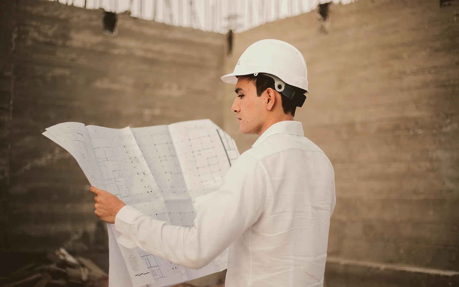Two contractors in work jackets lean over a folding table in an unfinished room, one pressing a marked up floor plan flat while the other points at a line on it