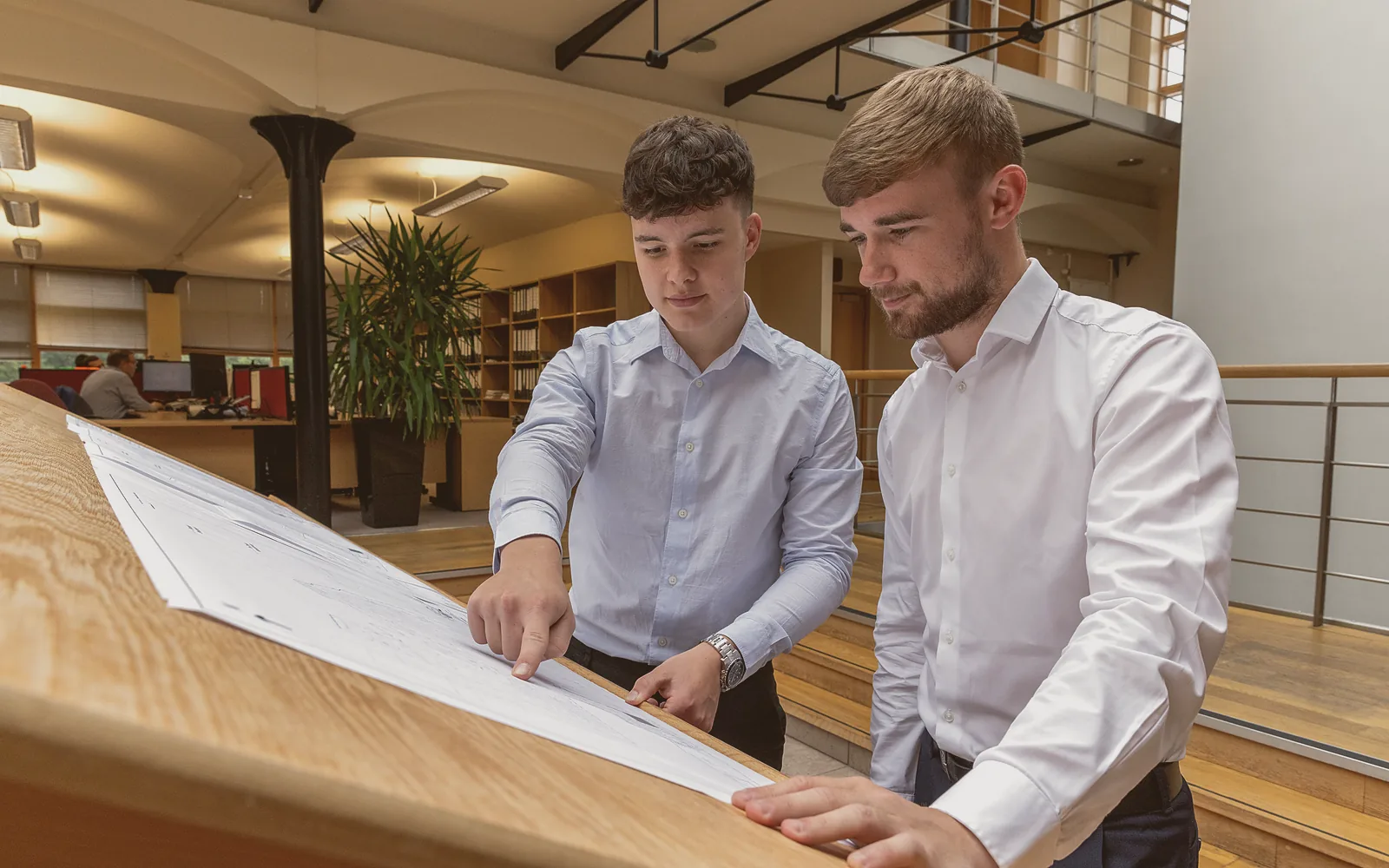 Two people at a desk in a small office, one at a laptop with a website open and the other pointing at the screen with a pen, daylight from a window