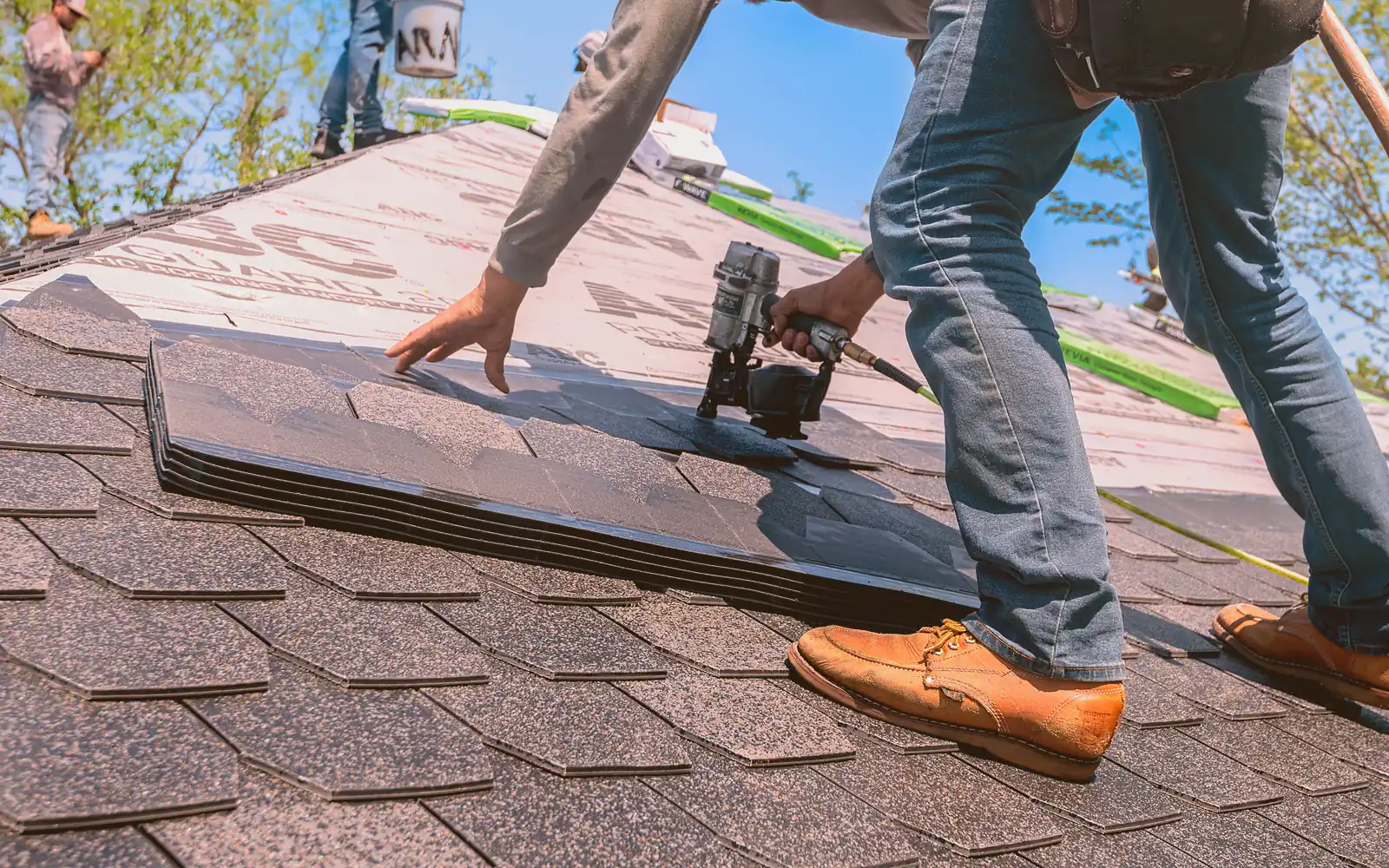 A roofing foreman in work clothes checks a tablet on a residential roof while two crew members lay shingles behind him