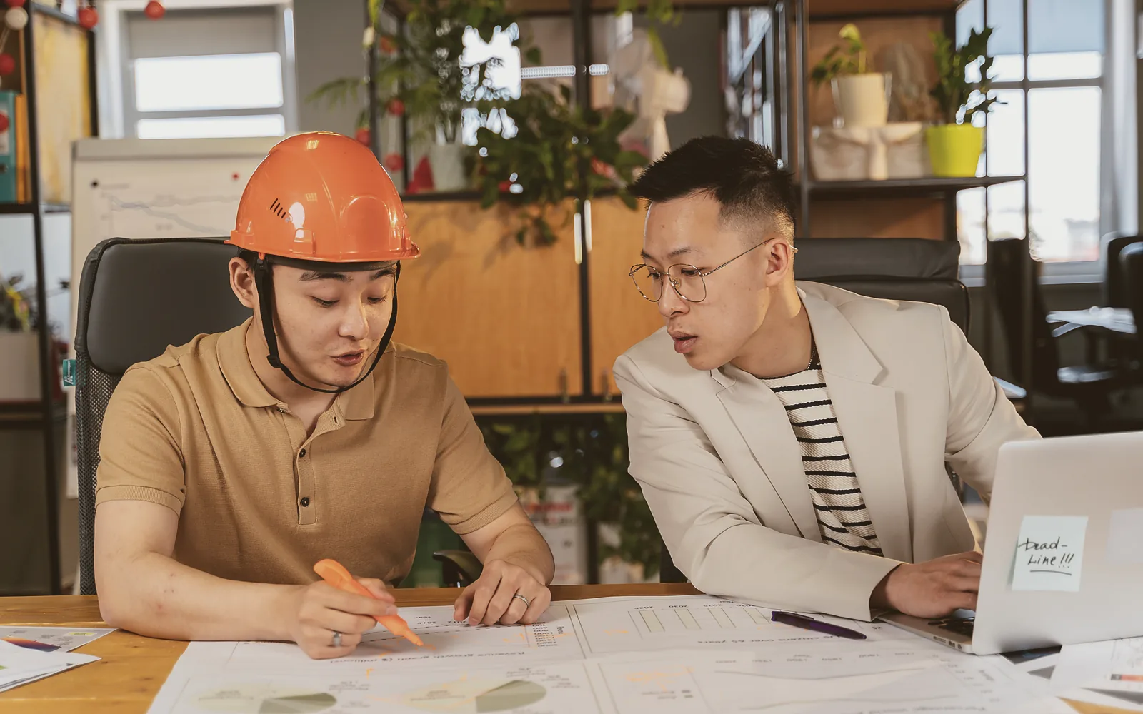 Two people in a small office lean over the same laptop on a cluttered desk while one of them holds a pen above a printed sheet
