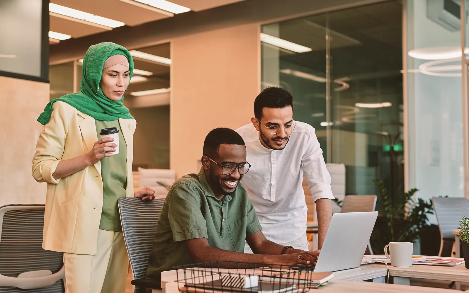 Two coworkers lean over the same laptop on an office desk while one of them holds a printed sheet