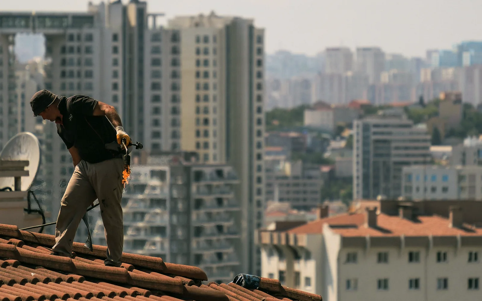 A roofer seals the roof he was hired for while a whole city of rooftops waits behind him