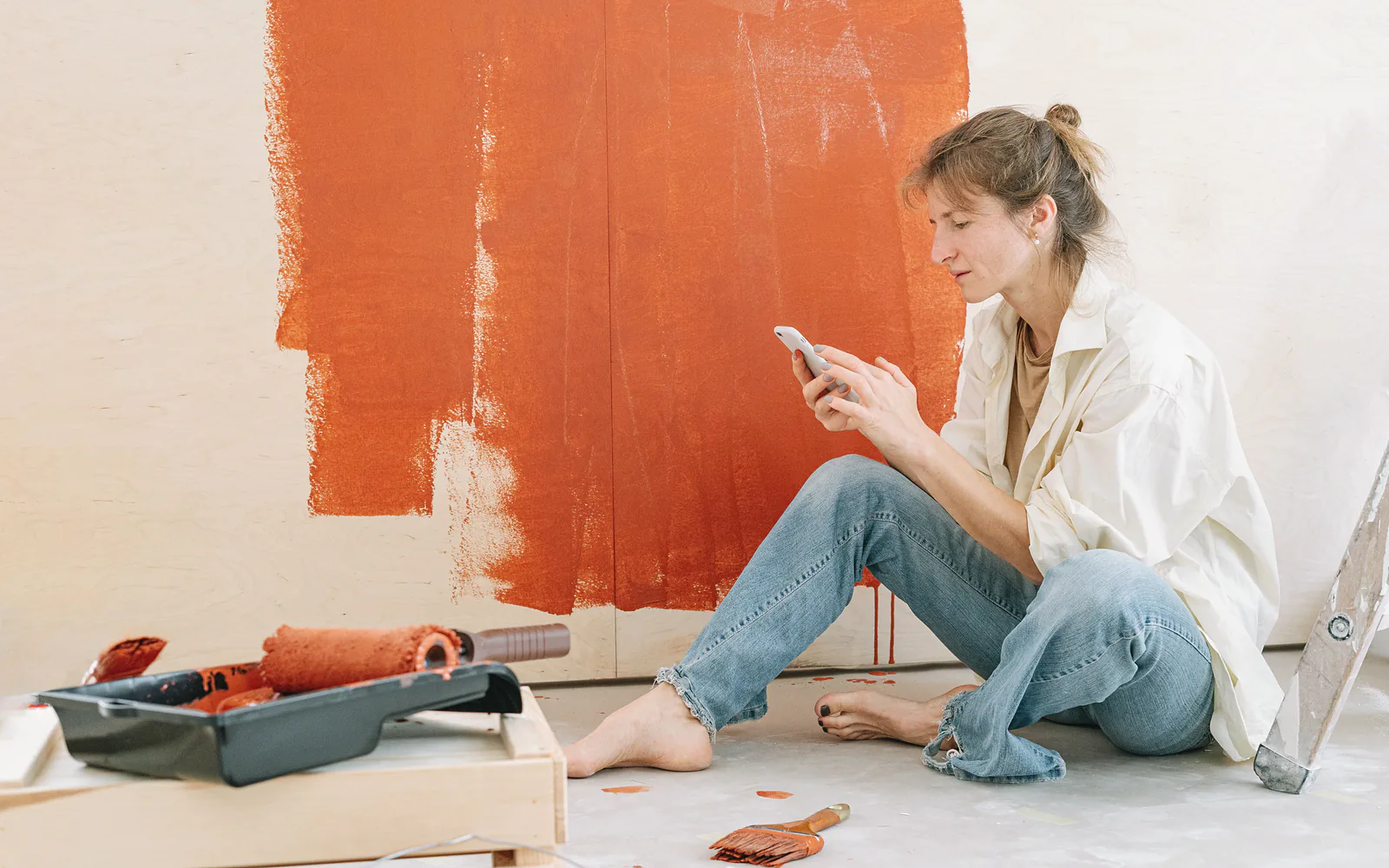 A woman sits on the floor of a half-painted room and checks her phone between coats
